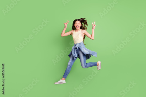 Full length body size view of pretty funky cheerful girl jumping walking showing v-sign isolated over bright green color background
