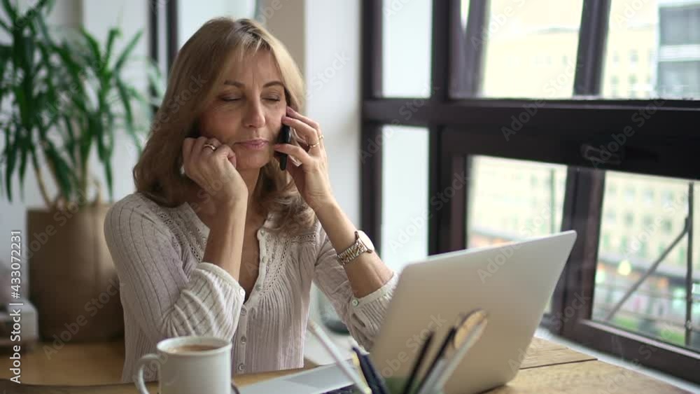Businesswoman talking on phone and using laptop at table in modern home office. Businesswoman talking on phone and using laptop at table in modern home office.