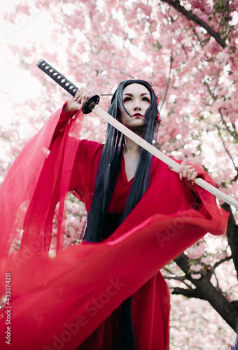 Young woman portrays geisha with sword in her hand near blooming sakura trees.
