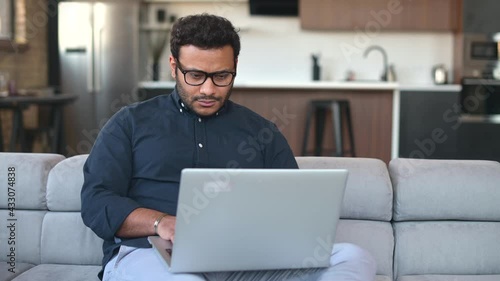 Serious multiracial man using laptop computer for remote work from home, hindu freelancer guy reading incoming emails, chatting online, researching or studying online sitting on the couch at home