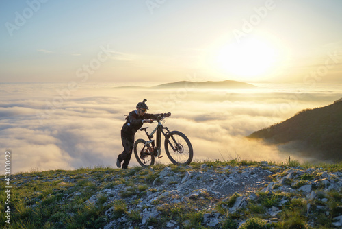 Mountain Biker Pushing Bike Up to the Mountain Above the Clouds at Sunset.
