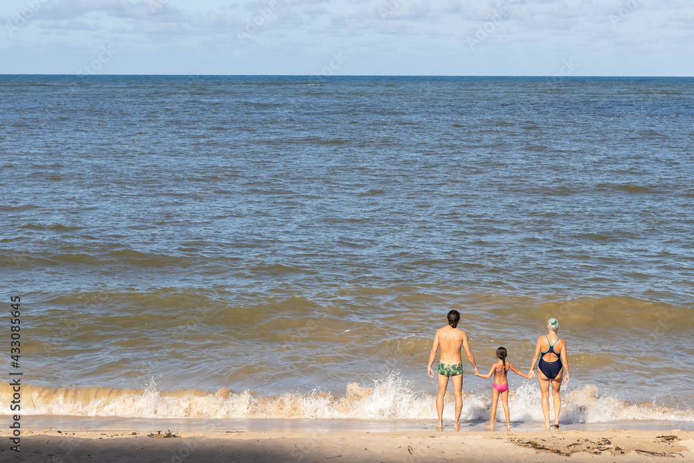 Homem adulto, criança e mulher idosa de mãos dadas e de frente para o mar Homem adulto, criança e mulher idosa de mãos dadas e de frente para o mar