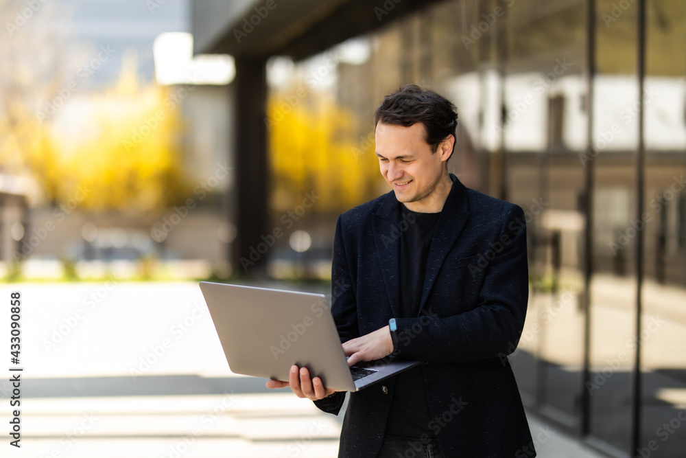 Portrait of caucasian businessman using and looking at laptop while standing outdoors near building Portrait of caucasian businessman using and looking at laptop while standing outdoors near building