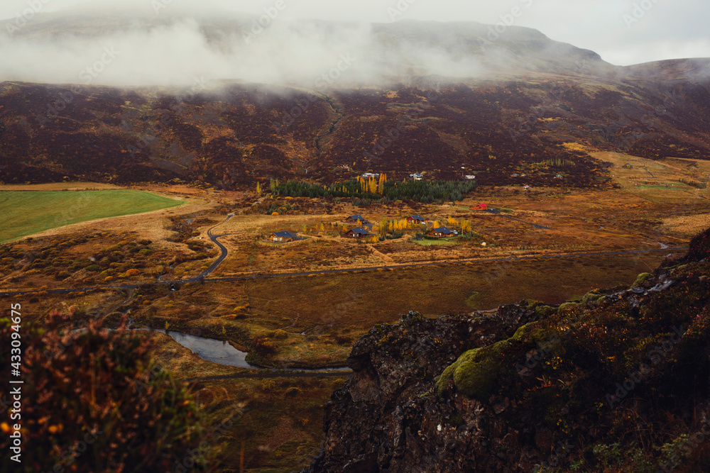 Aussicht eines Wanderers vom Gipfel eines Berges auf ein Dorf im Tal durchzogen von einem Bach, Wiesen und Weiden und tief hängenden Wolken und kondensierendes Wasser beim Wandern