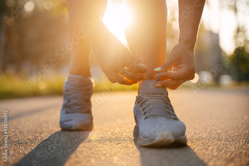 Running woman tying the laces of sneakers before jogging on the road in the park in sunny weather, training in nature.