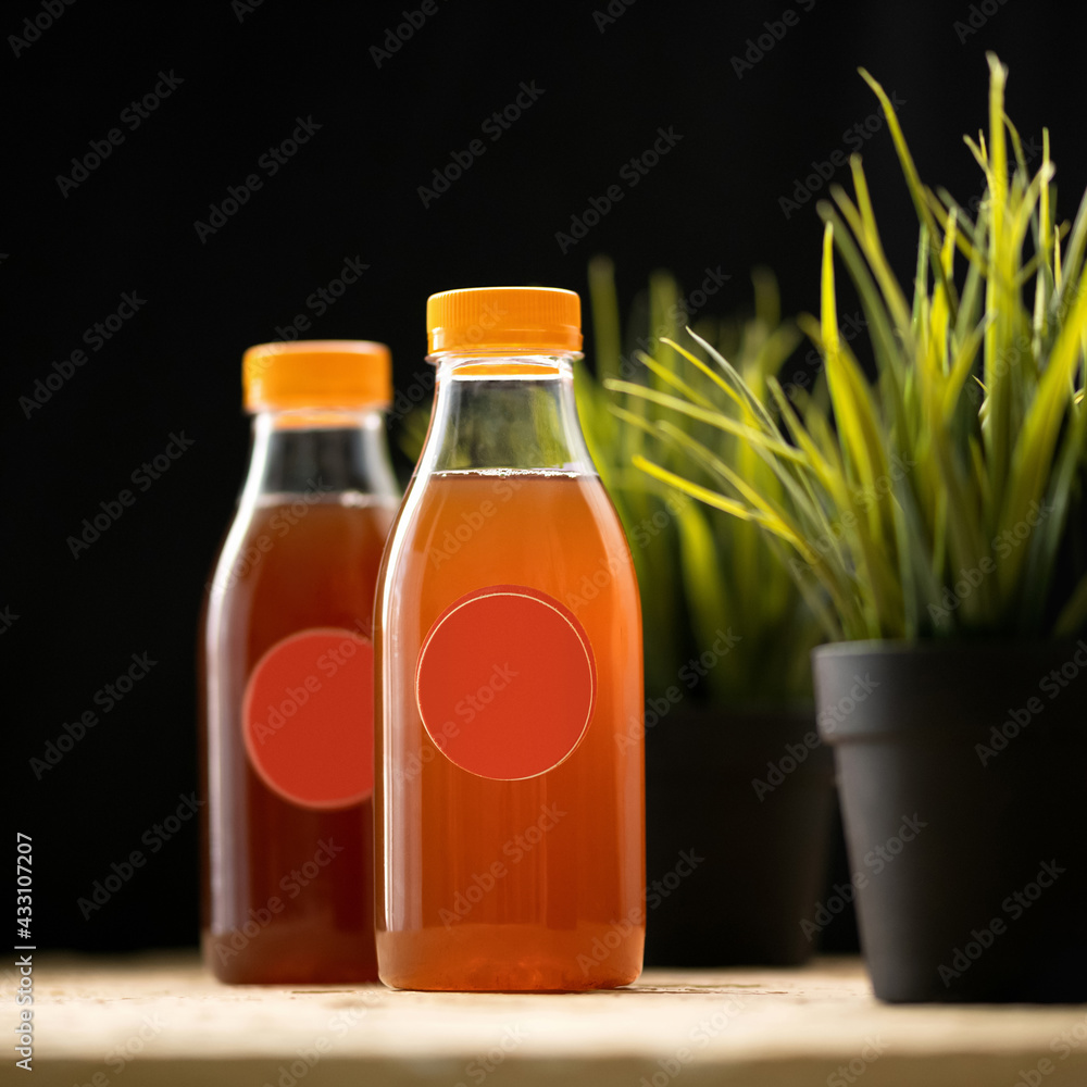 Blank label at transparent bottles with orange juice or soft drink at table with artificial flower pots on it. Black background. Bright colors of nature. Mock up or template for label design. Blank label at transparent bottles with orange juice or soft drink at table with artificial flower pots on it. Black background. Bright colors of nature. Mock up or template for label design.