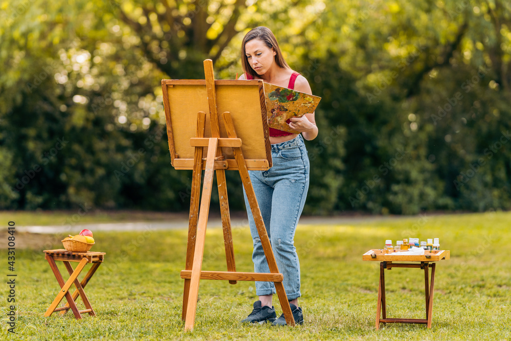 Woman painting on canvas in a park with two side tables with tools Woman painting on canvas in a park with two side tables with tools
