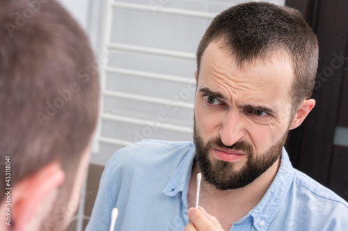Man cleans his ears with cotton swabs, person looking himself in bathroom mirror, portrait, close-up