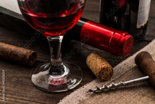 Two bottles of red wine with a glass of red wine and cuban cigar with corkscrew on an old wooden table. Close up view, focus on the glass of red wine