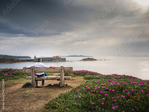 man relaxes sitting on a wooden bench looking at the sea surrounded by beautiful greenery on a spring afternoon