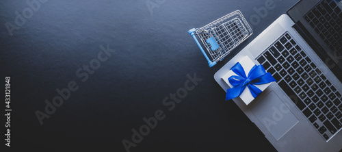 Online shopping background. Laptop computer, shopping trolley and white gift with blue ribbon on dark. Internet purchase, online shop concept. Website retail business.