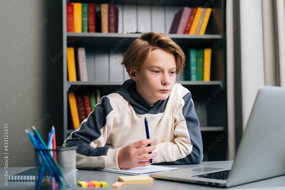 Close-up of thinking pupil boy thoughtful writing in notebook with pen sitting at desk near window. Shotting from below of child schoolboy doing homework at home. Close-up of thinking pupil boy thoughtful writing in notebook with pen sitting at desk near window. Shotting from below of child schoolboy doing homework at home.