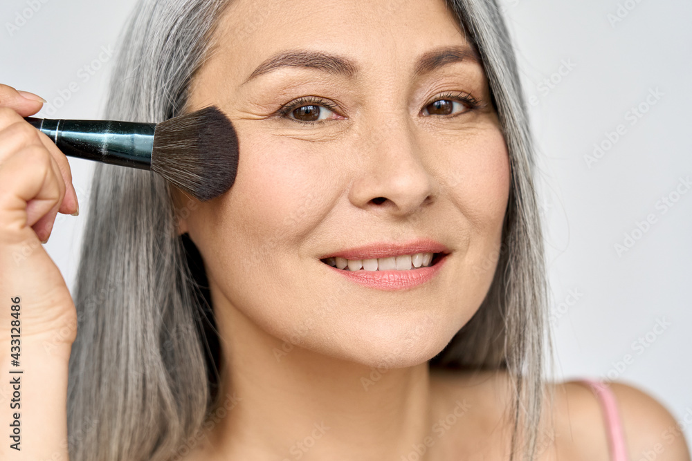 Senior older middle aged Asian woman with grey hair holding make up brush  on radiant face