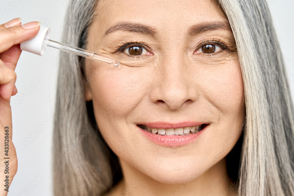 Closeup cut portrait of senior mature older Asian woman touching clean face eye contour with antiaging pipette serum essence oil looking at camera. Anti wrinkle prevention skin care products concept.