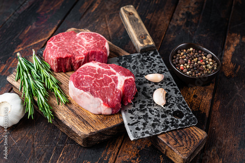 Fresh Raw Fillet Mignon tenderloin steaks on butcher cutting board with meat cleaver. Dark wooden background. Top view