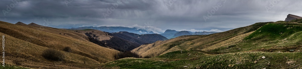 Panoramic landscape view over Gum-bashi mountain pass, with new green and old brown grass on cliffs, dark clouds before the storm, Сaucasus mountian range and Elbrus mountain. Karachay-Cherkessia