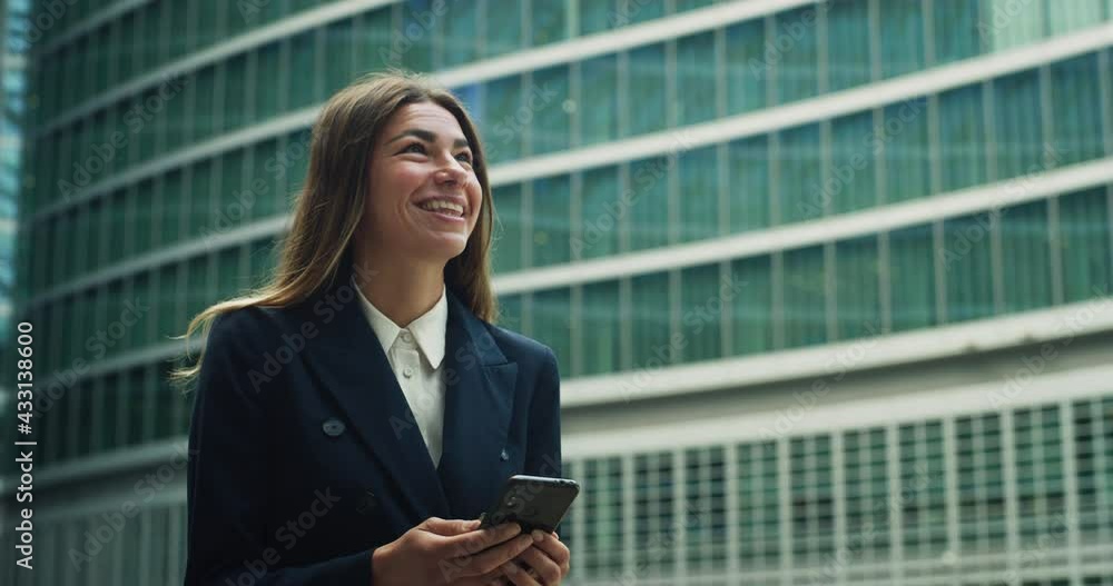 Cinematic shot of smiling business woman using smartphone for sending emails and make calls for work on skyscrapers background. Concept of technology, network, connection, corporate, finance, success. Cinematic shot of smiling business woman using smartphone for sending emails and make calls for work on skyscrapers background. Concept of technology, network, connection, corporate, finance, success.
