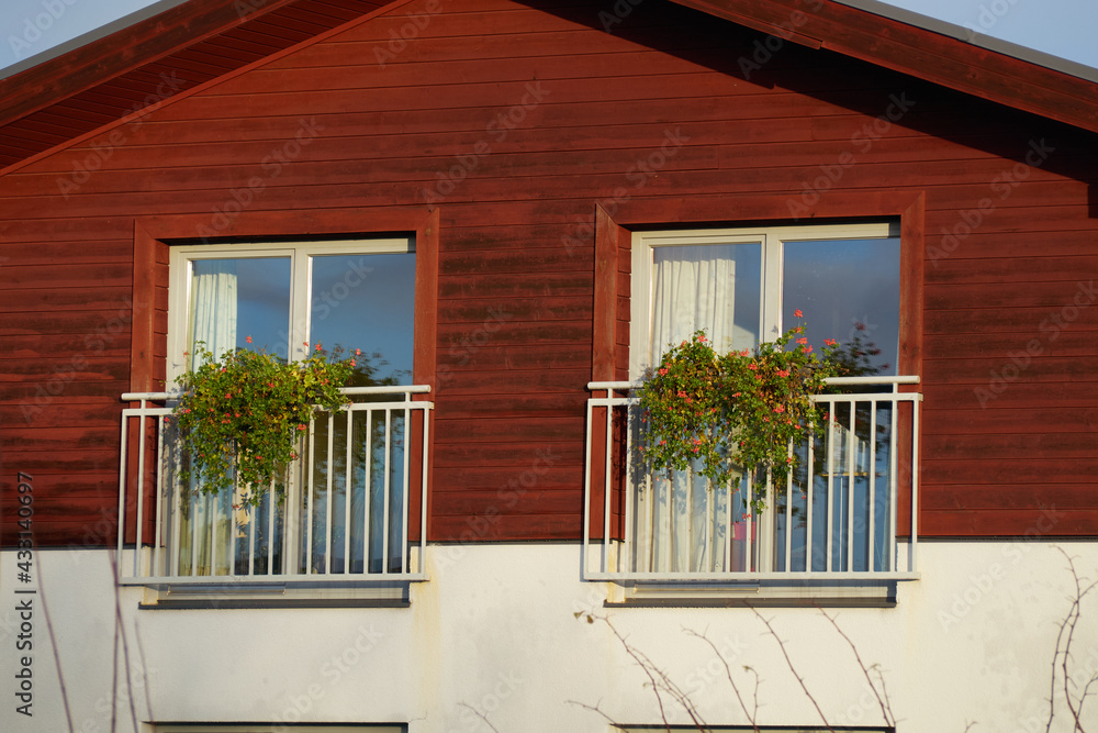 Two plastic windows with plants on balcony and red wooden house wall.