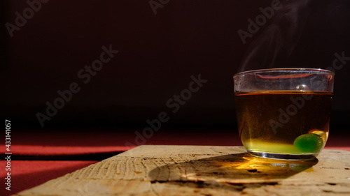 Fully linden tea filled glass pot existing on wooden plate and red carpet with black shadow background. Isolated linden tea cup made of glass and there is green mentholated sweat and candy.  photo