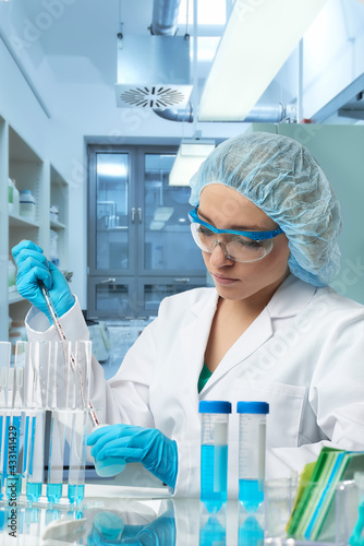 Fototapeta Naklejka Na Ścianę i Meble -  Pharma female tech works in laboratory. Caucasian, european woman, young adult in protective gloves, hat and white gown handles sample tube. Defocused lab interior, selective focus. Biotech research.