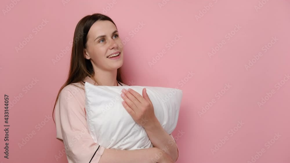 Portrait of dreamful cute young woman wearing pajama standing over pink isolated background looking away to side with smile on face, natural expression, holding white pillow. Laughing confident. Portrait of dreamful cute young woman wearing pajama standing over pink isolated background looking away to side with smile on face, natural expression, holding white pillow. Laughing confident.
