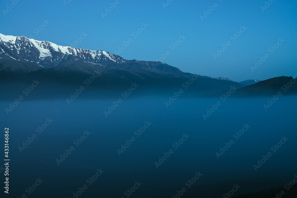 Atmospheric mountains landscape with dense fog and great snow mountain range under twilight sky. Alpine scenery with big snowy mountain ridge over thick fog in night. Snowy rocks above clouds in dusk.
