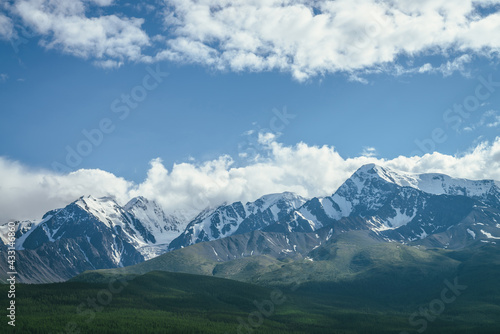 Awesome mountains landscape with sunlit high snowy pinnacle among low clouds in blue sky. Atmospheric highland scenery with snow-white big mountain top in sunlight. Wonderful snowy mountain range.