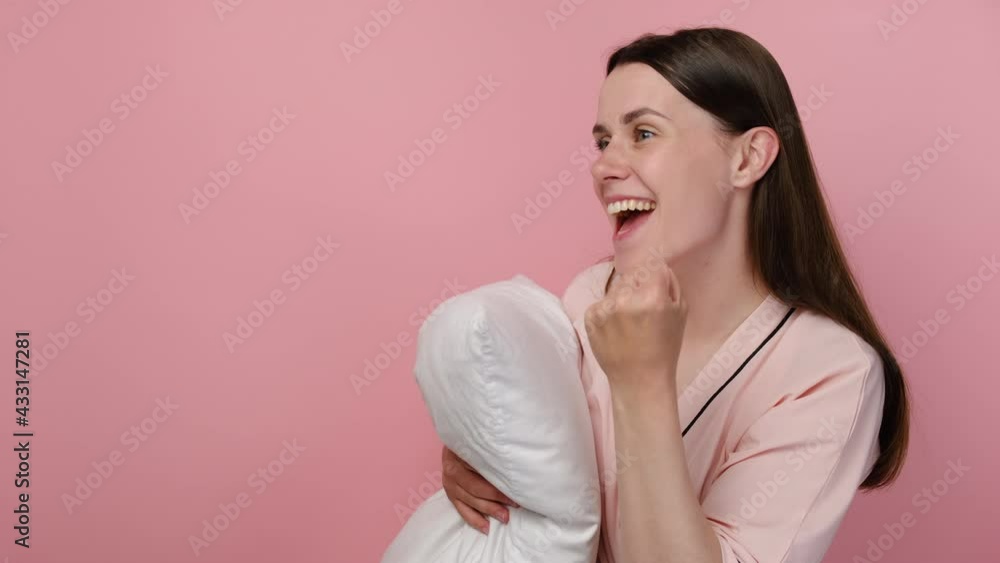 Side view of shocked excited young woman wearing pajama standing over pink isolated background celebrating surprised and amazed for success with arms raised, holding white pillow. Winner concept Side view of shocked excited young woman wearing pajama standing over pink isolated background celebrating surprised and amazed for success with arms raised, holding white pillow. Winner concept