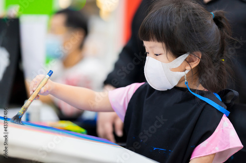 Cute girl wearing a 3D surgical face mask is taking art classes in classroom. Child make crafts with brushes painting water color on a canvas. Children wear black apron uniforms. Kid is 4 years old.