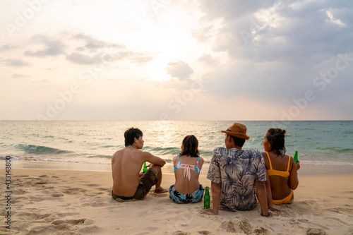Group of Happy Asian man and woman friends sitting on the beach enjoy drinking beer with talking together at summer sunset. Male and female friendship relax and having fun outdoor lifestyle activity.