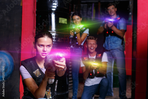 Portrait of young positive cheerful smiling girl took aim colored laser guns during laser tag game in labyrinth