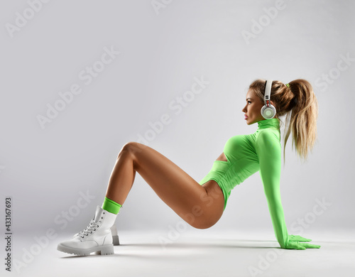 Slim blonde woman fitness trainer in stylish sportswear bodysuit and brutal shoes does pelvic lifts on floor, standing in bridge position and listen music over light background photo