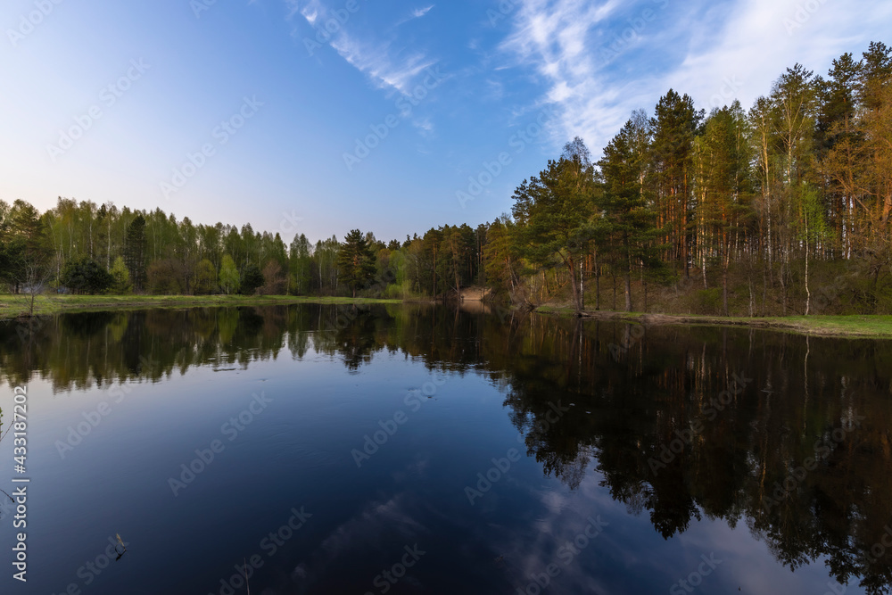 Spring landscape, river, bushes and trees, reflected in the water. The blue sky is reflected in the water. The rays of the setting sun illuminate the trees.