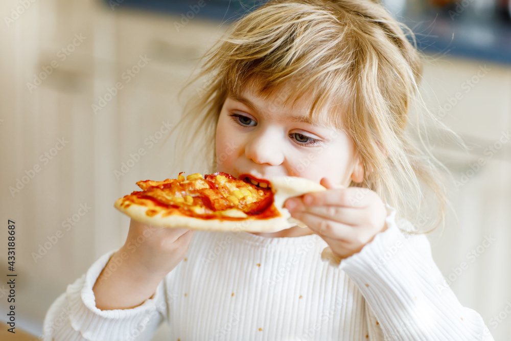 Adorable toddler girl eat italian pizza with vegetables and chees. Happy child eating fresh cooked healthy meal with tomatoes, corn and vegetables at home, indoors.
