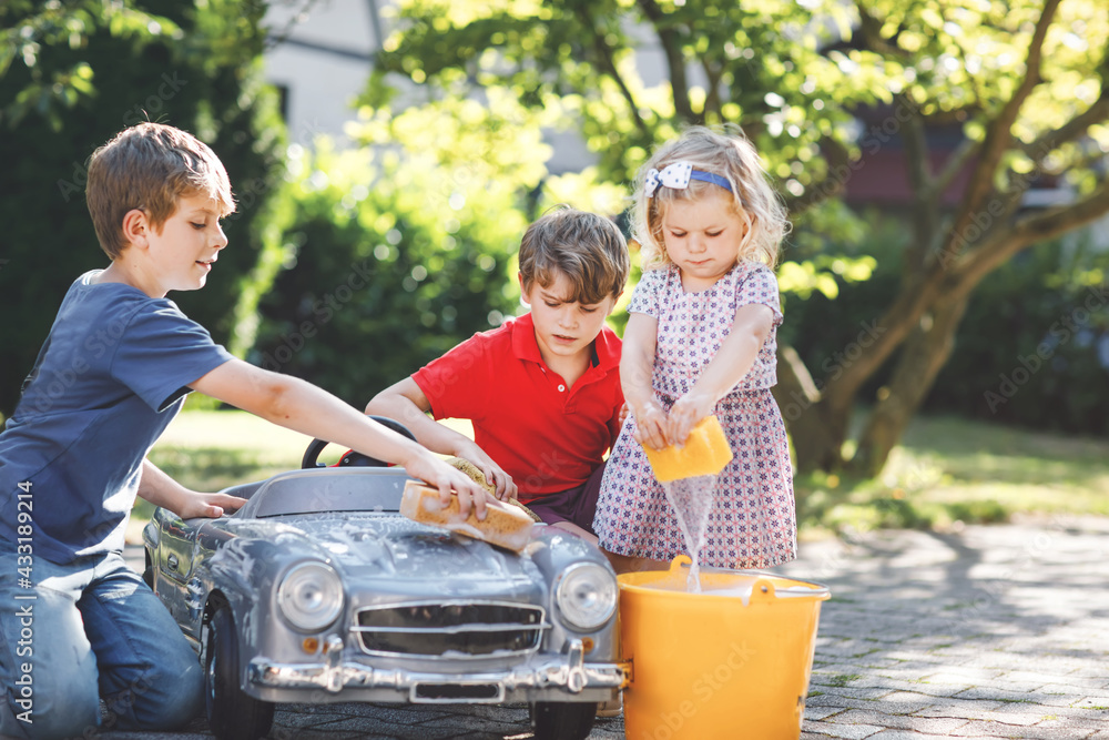 Three happy children washing big old toy car in summer garden, outdoors. Two boys and little toddler girl cleaning car with soap and water, having fun with splashing and playing with sponge.