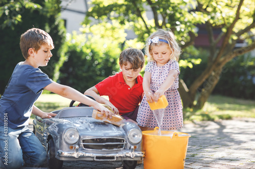 Three happy children washing big old toy car in summer garden, outdoors. Two boys and little toddler girl cleaning car with soap and water, having fun with splashing and playing with sponge. © Irina Schmidt