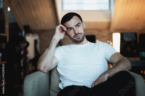 Portrait of handsome serious young brunette man in white t-shirt sitting in armchair at home