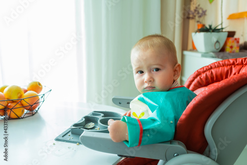 A serious dirty-faced baby boy of 8 monthes is eating with hands at the table. He is sitting in a high chair at the white table with his plate on it. Baby-led weaning concept. photo