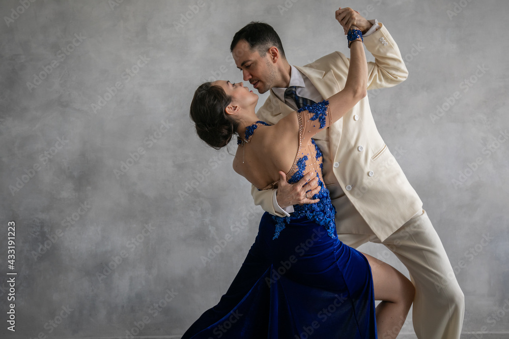 Couple of professional tango dancers in elegant suit and blue dress pose in a dancing movement on grey background. Handsome man and woman dance looking eye to eye. - obrazy, fototapety, plakaty 