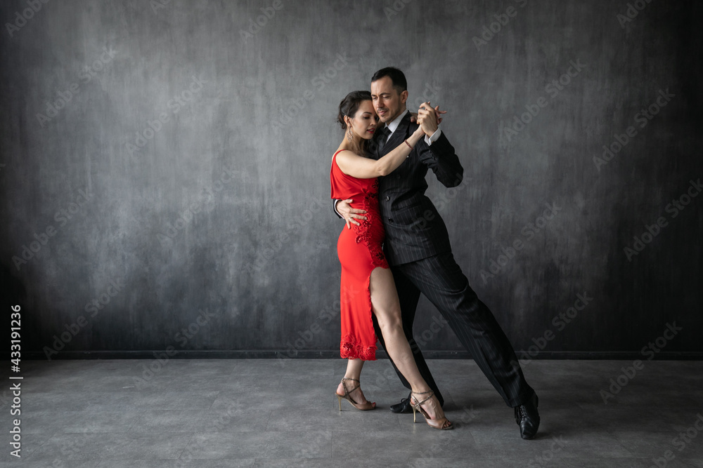 Couple of professional dancers in elegant suit and red dress in a tango dancing movement on dark background. Handsome man and woman dance cheek to cheek. - obrazy, fototapety, plakaty 