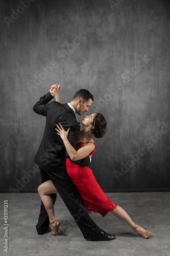 Couple of professional dancers in elegant suit and red dress in a tango dancing movement on dark background. Handsome man and woman dance looking  eye to eye. photo