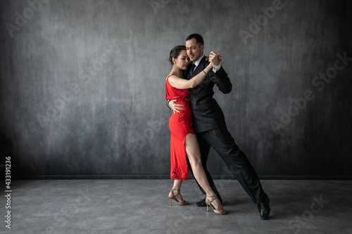 Couple of professional dancers in elegant suit and red dress in a tango dancing movement on dark background. Handsome man and woman dance cheek to cheek.