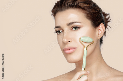 Beautiful woman with fresh clean skin massaging face with jade roller. Conceptual of skin care and beauty facial treatments with semi precious stones. Close up face isolated on beige with copy space
