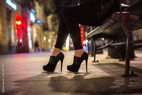 High-heeled legs of a girl sitting on a bench at night against the blurred city lights