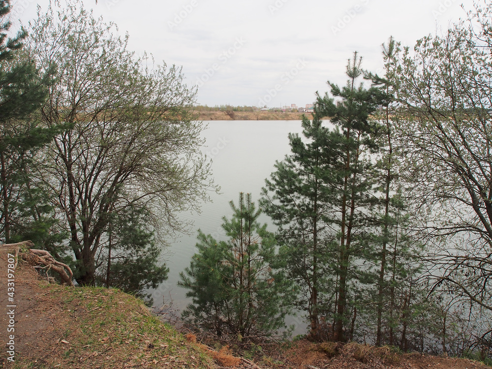 Spring landscape with pine trees and deciduous trees on the steep shore of the lake, against the background of a gloomy sky. Rural landscape, a beautiful place to relax. Spring landscape with pine trees and deciduous trees on the steep shore of the lake, against the background of a gloomy sky. Rural landscape, a beautiful place to relax.