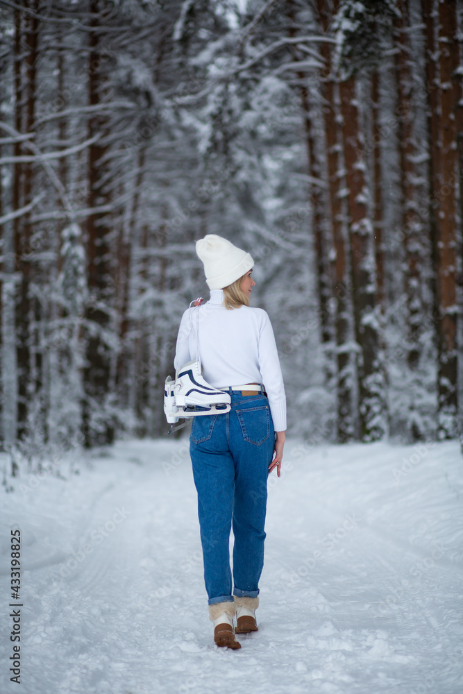 Young blond beautiful female with white ice skates in her hand in winter snowy forest.