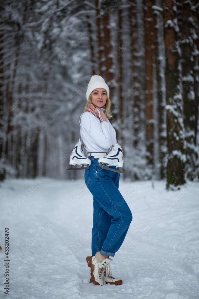 Young blond beautiful female with white ice skates in her hand in winter snowy forest.