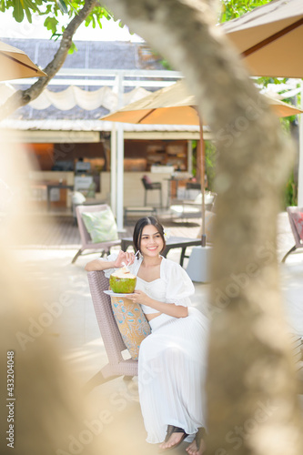 Beautiful woman tourist with white flower on her hair drinking coconut sitting on lounge chair during summer holidays