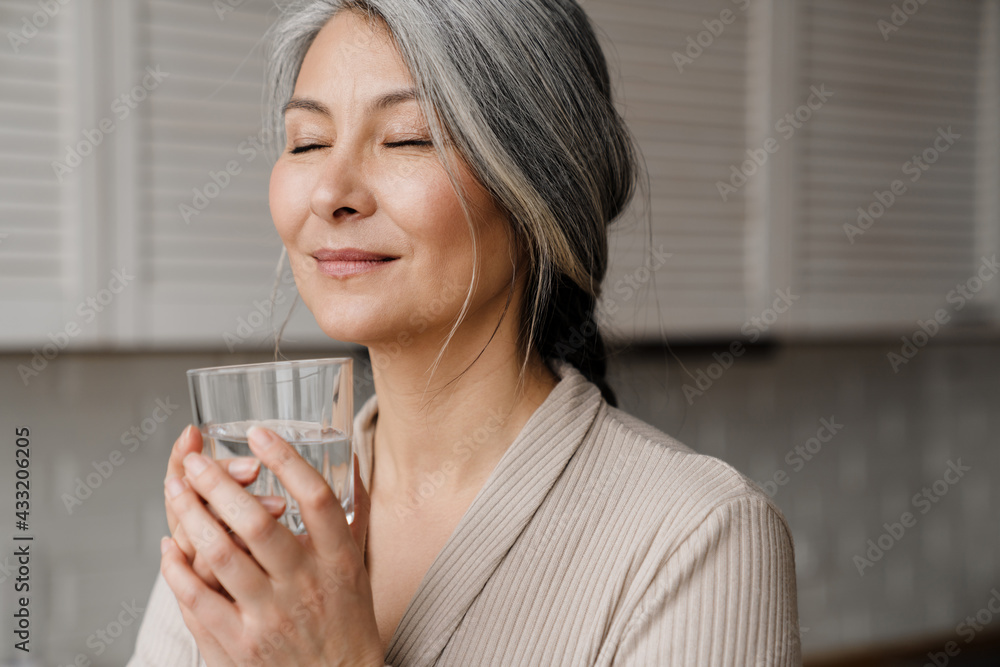 Mature grey woman smiling while drinking water in kitchen at home Mature grey woman smiling while drinking water in kitchen at home
