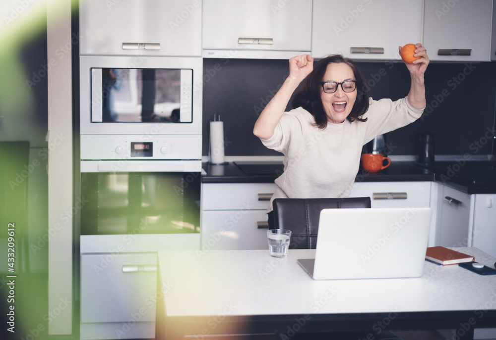 Beautiful happy mature elderly woman with orange in her hands standing at home in the kitchen in front of laptop screen, healthy eating and active retirees concept Beautiful happy mature elderly woman with orange in her hands standing at home in the kitchen in front of laptop screen, healthy eating and active retirees concept